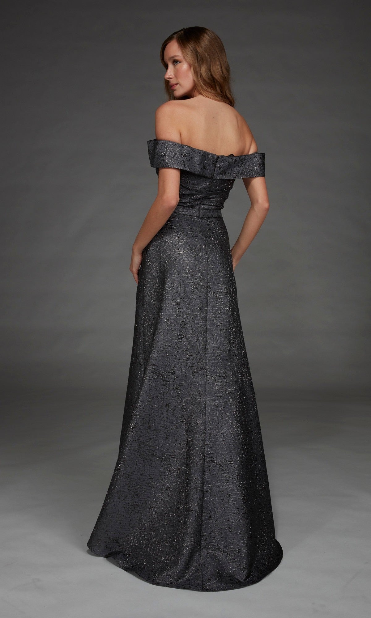 A woman models the Alyce 27882 long formal dress, featuring an off-the-shoulder jacquard design, as she stands facing away and looks over her shoulder against a plain gray background.